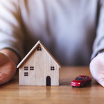 Figures of a wooden home and a red car between a person's hands. The figures are on a brown table.