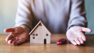 Figures of a wooden home and a red car between a person's hands. The figures are on a brown table.